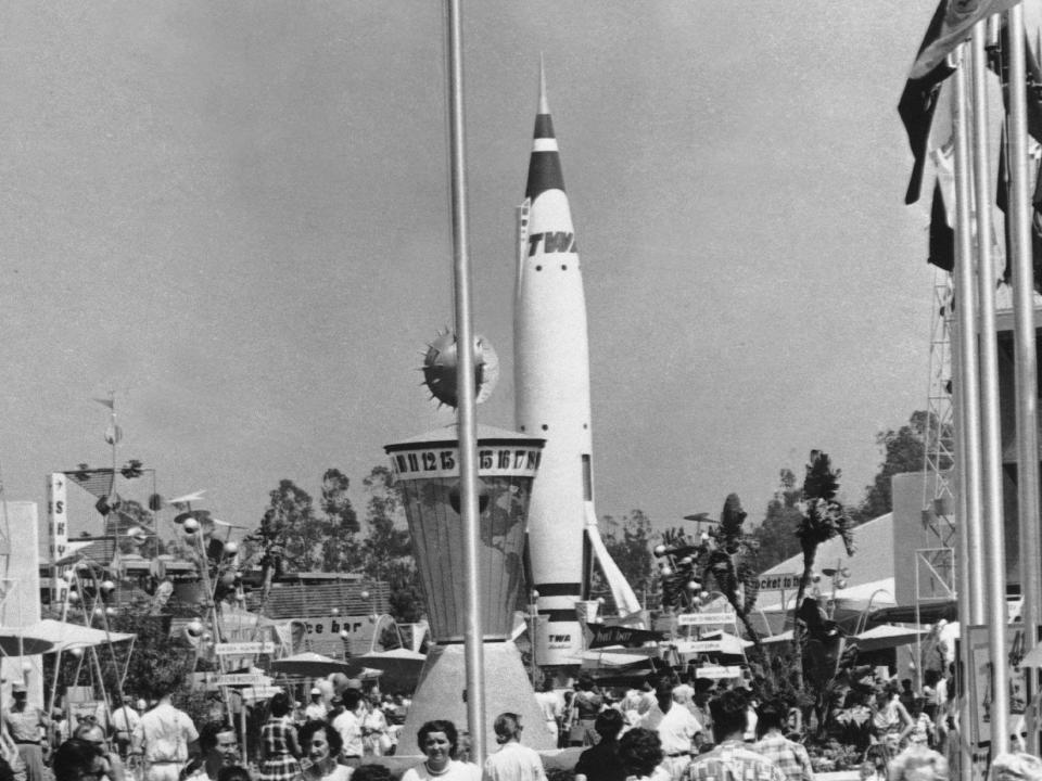 Black and white photo from the entrance of the theme park with many people and a tall rocketship.