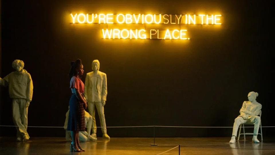 Art gallery director Brianna Cartwright (Teyonah Parris) walks across an exhibit, showing three haphazard white mannequins with neon letters hanging over which read "You're Obviously in the Wrong Place."