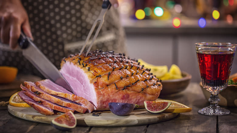 Person slicing a glazed ham beside a glass of red wine