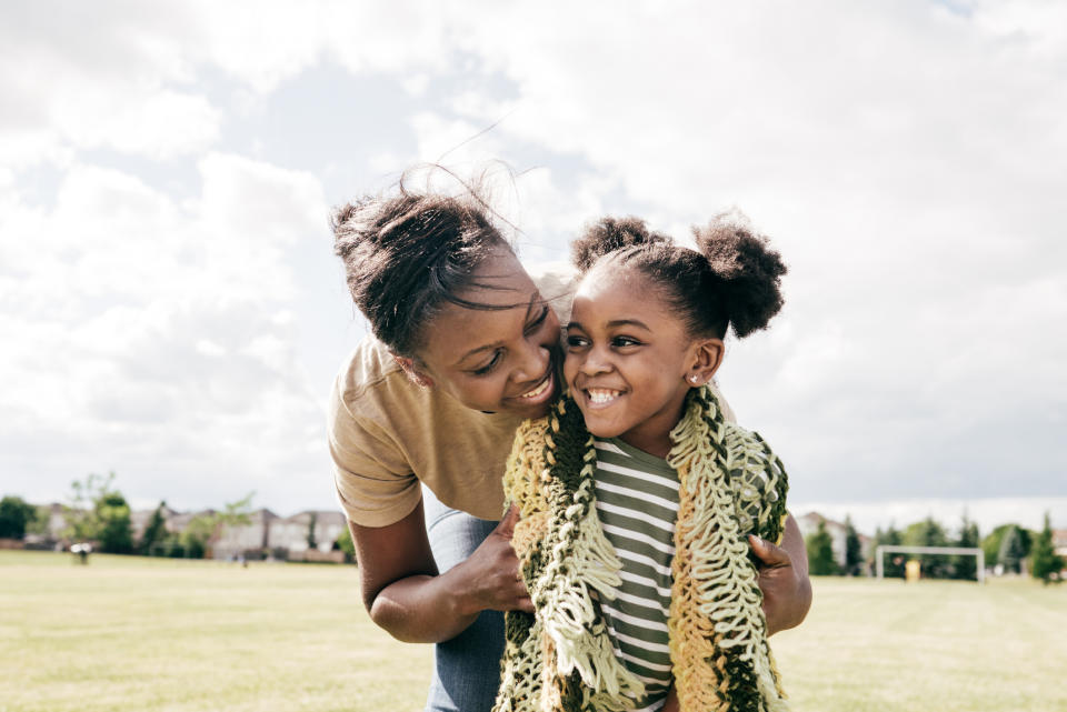 A mother joyfully embraces her smiling daughter in a park. The mother wears a short-sleeved top, while the daughter wears a striped shirt and a knitted scarf