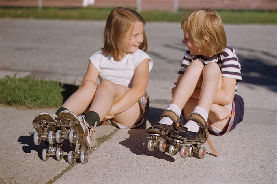 Two young girls, seated on a sidewalk, laughing and wearing roller skates