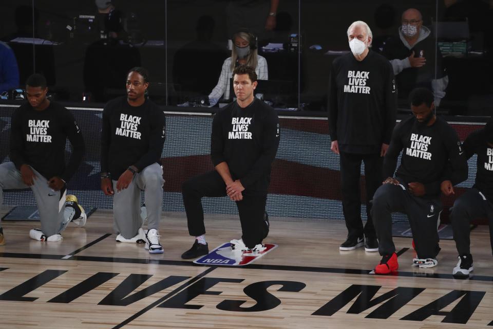 San Antonio Spurs head coach Gregg Popovich, fourth from left, stands while wearing a mask while Sacramento Kings head coach Luke Walton, center, kneels with players before an NBA basketball game Friday, July 31, 2020, in Lake Buena Vista, Fla. (Kim Klement/Pool Photo via AP)