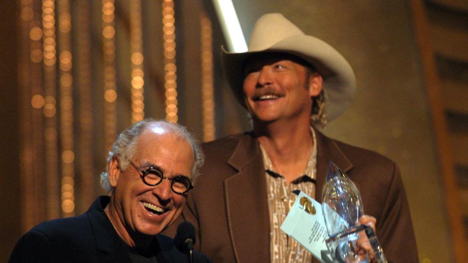 jimmy buffett smiles and stands in front of a microphone holding a glass award, behind him stands alan jackson in a cowboy hat