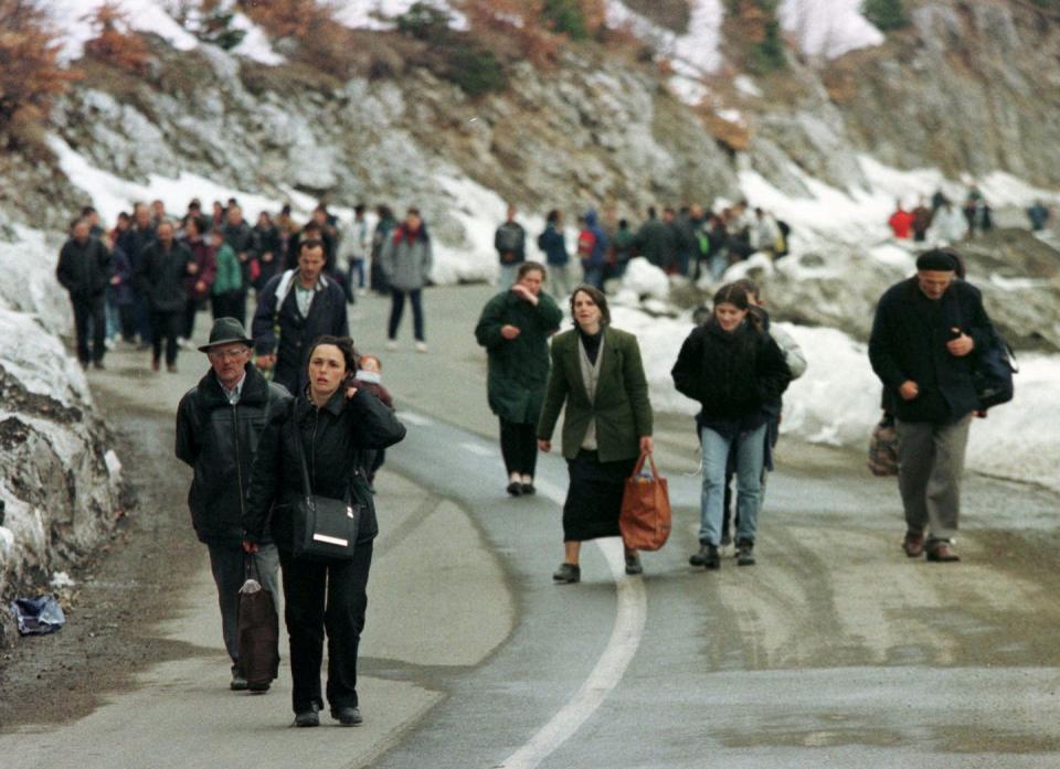 People walk on a mountainous road, wearing backpacks and carrying language; there is snow on the ground