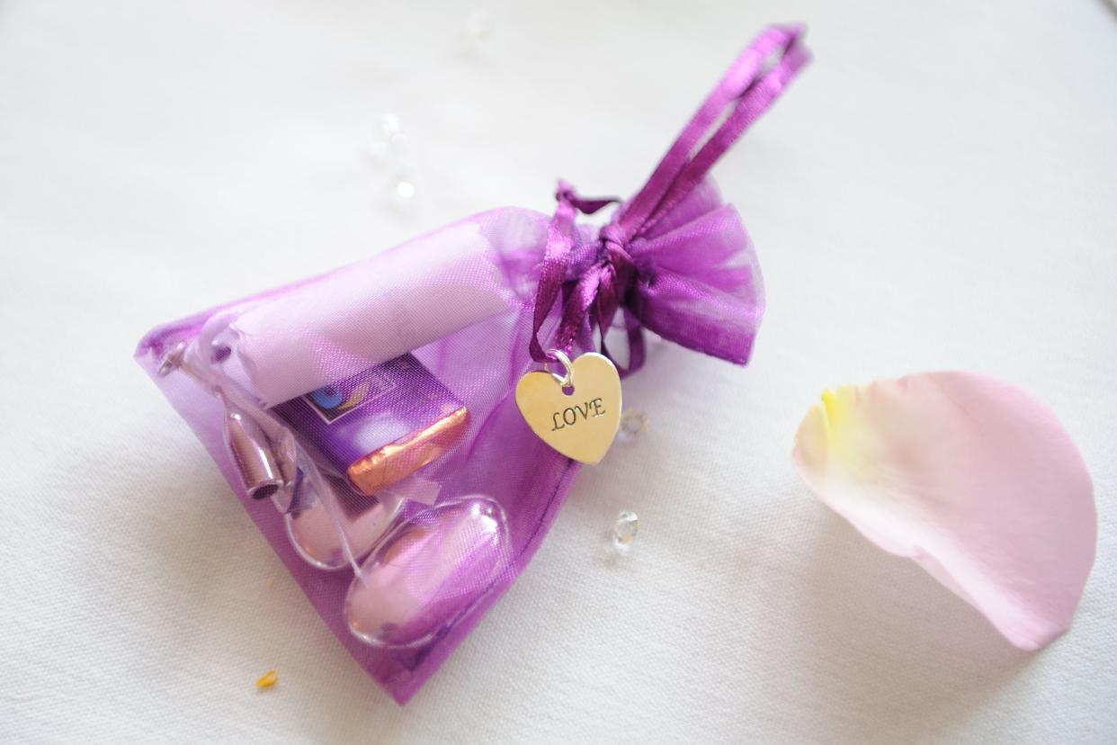 Closeup of a delicate purple wedding gift bag, see-through with a piece of chocolate and a tiny champagne glass trinket, next to one lavender colored rose petal on a light blue background