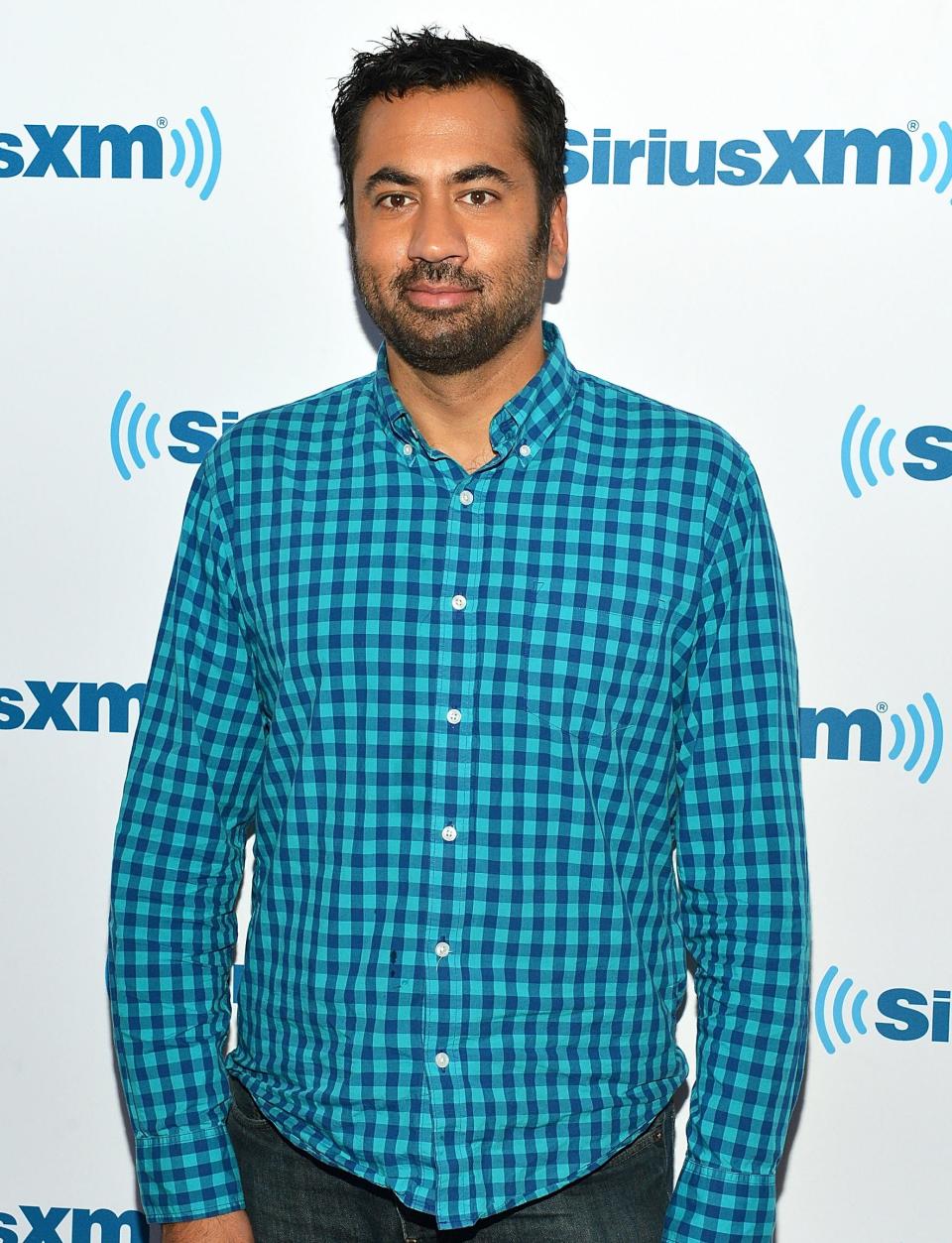 Kal Penn in checkered shirt and jeans at a SiriusXM event backdrop