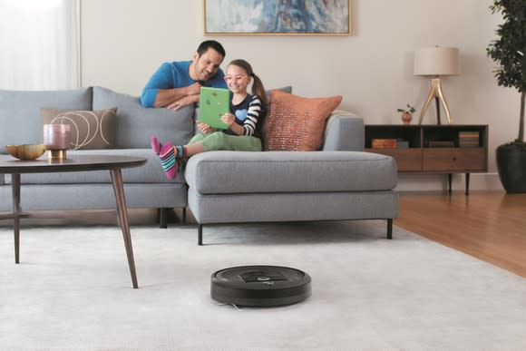 A man and a young girl looking at a tablet as she sits on a couch, with a robotic vacuum cleans the carpet in the foreground.