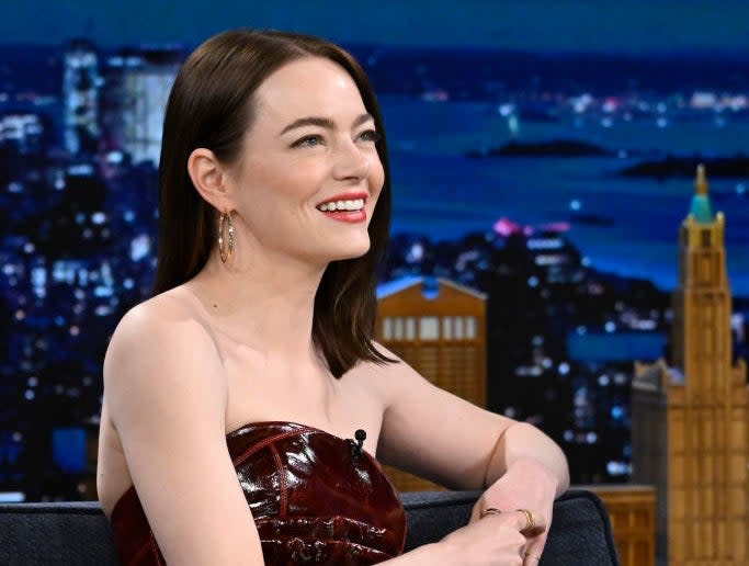 Emma Stone sits on a talk show set, smiling and wearing a strapless, glossy dress, during a nighttime city backdrop