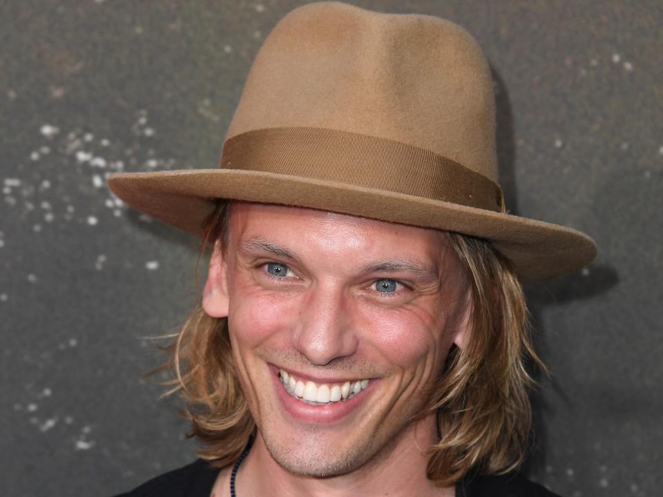 jamie campbell bower smiling on a red carpet, wearing a beige colored fedora over chin-length blonde hair, and a black jacket over a white t-shirt