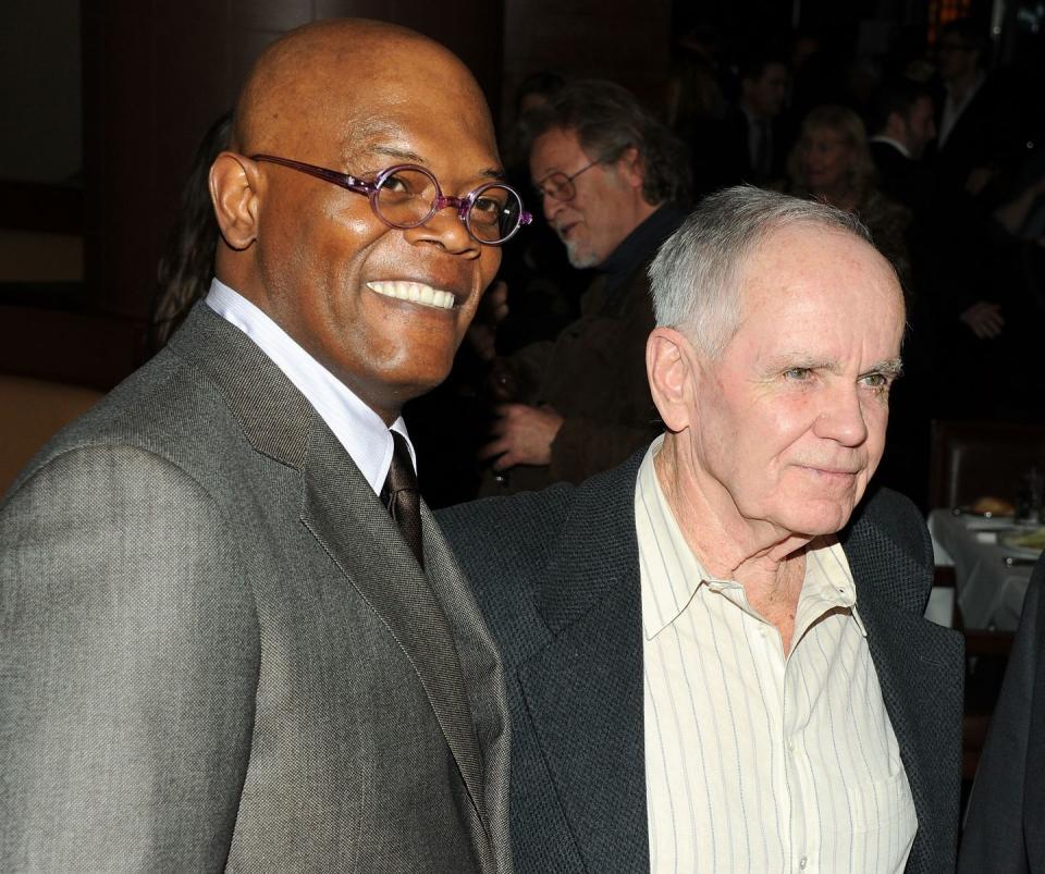 samuel l jackson, wearing a gray suit, black tie, and glasses, smiles as he stands next to cormac mccarthy, who wears a black suit jacket and white striped shirt