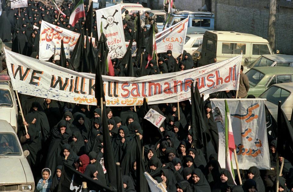 A crowd of people dressed in black protest and hold signs reading "we will kill salman rushdie"