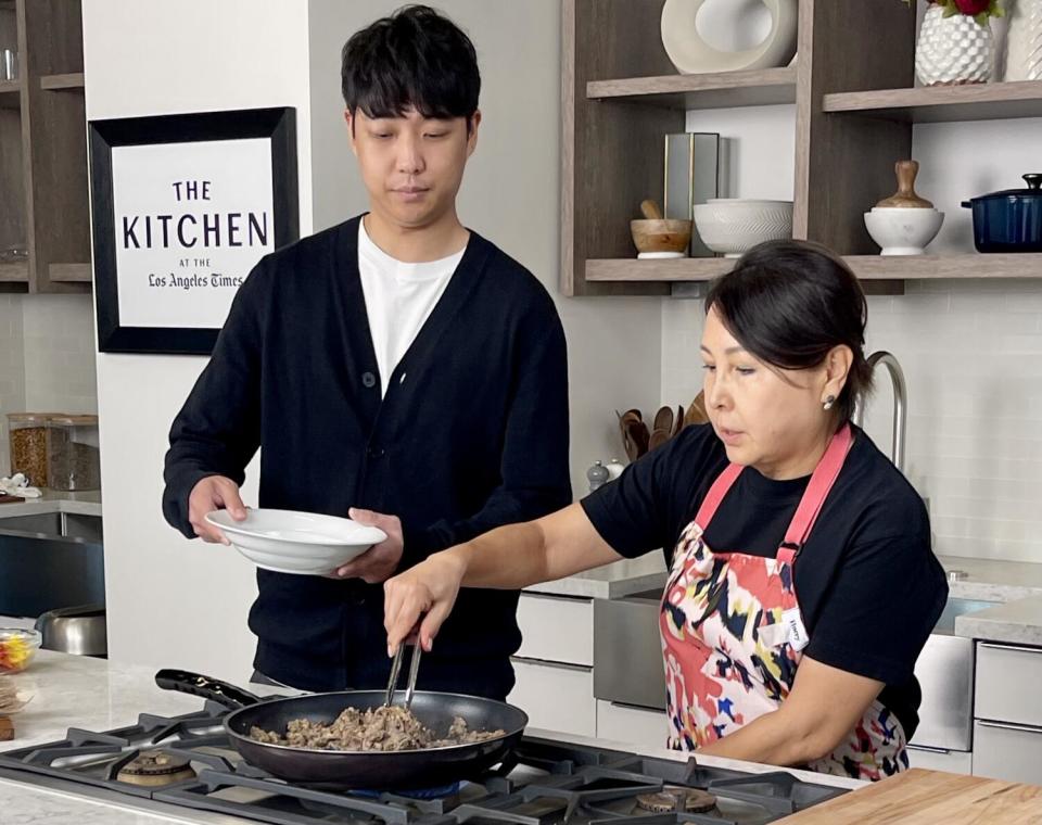 A young man and his mother cook meat in a skillet on a stovetop