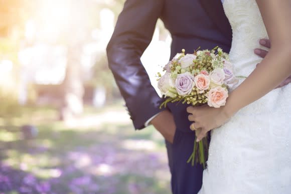 A couple in wedding outfits embraces while the woman carries a bouquet.