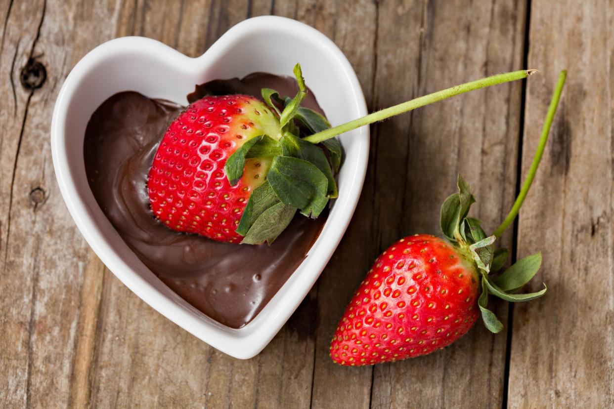 An overhead close up shot of a ripe red strawberry being dipped into a white heart shaped bowl full of melted chocolate. Shot on a grungy old wooden table.