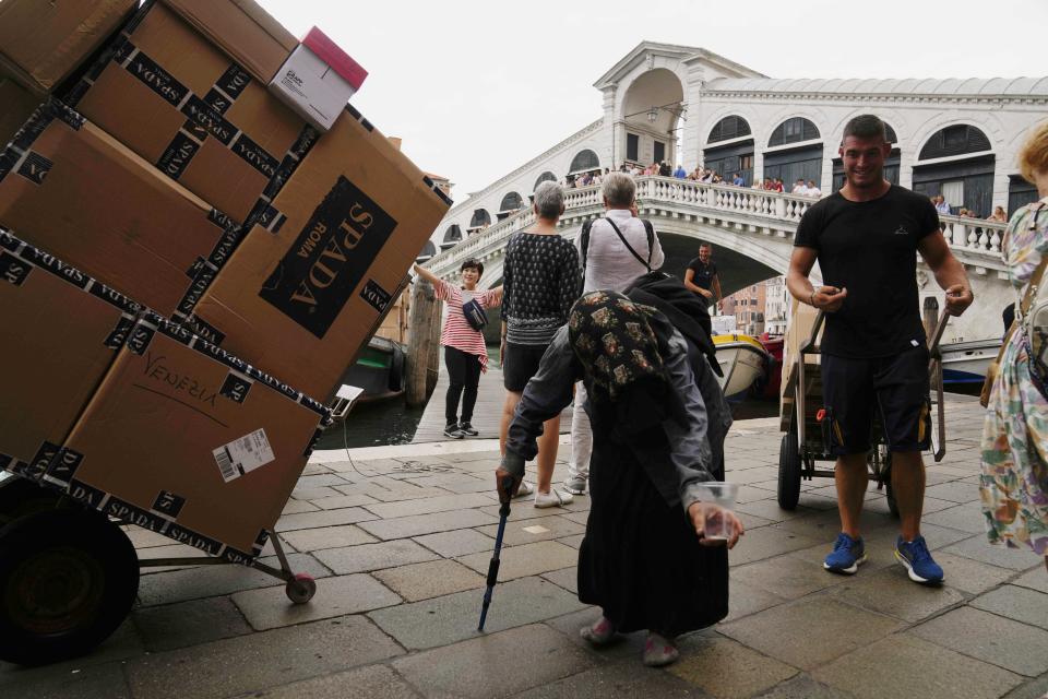 An old woman begs as workers carry supplies at the Rialto bridge in Venice, Italy, Wednesday, Sept. 13, 2023. The Italian city of Venice has been struggling to manage an onslaught of tourists in the budget travel era. The stakes for the fragile lagoon city are high this week as a UNESCO committee decides whether to insert Venice on its list of endangered sites. (AP Photo/Luca Bruno)