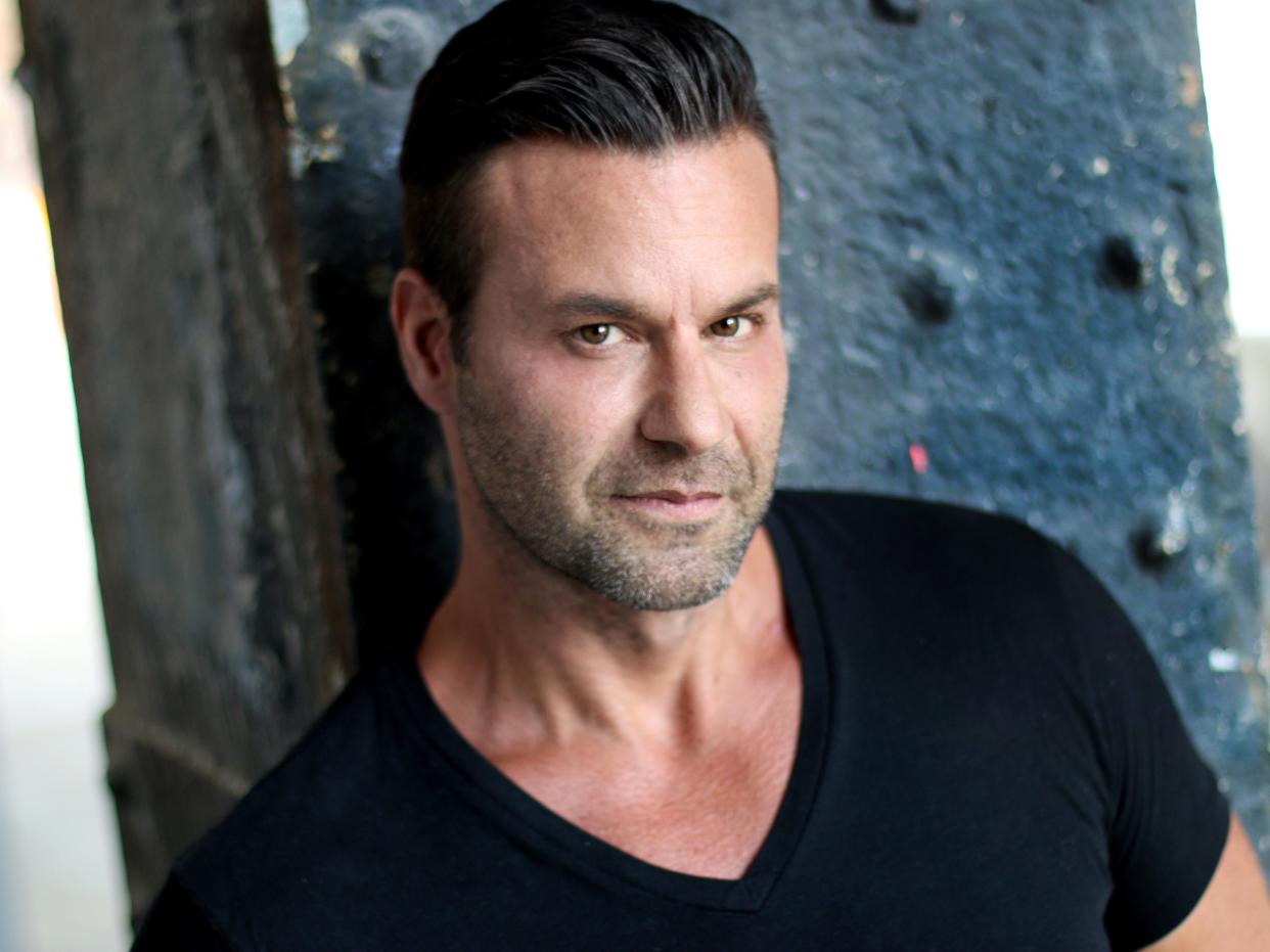 steve stanulis, a man with stubble, slicked back hair, a v-cut black tshirt leaning against a blue metal beam and looking upwards towards the camera.