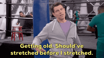 Max Greenfield in a gym, rubbing his back and saying, "Getting old. Should've stretched before I stretched." People are working out in the background