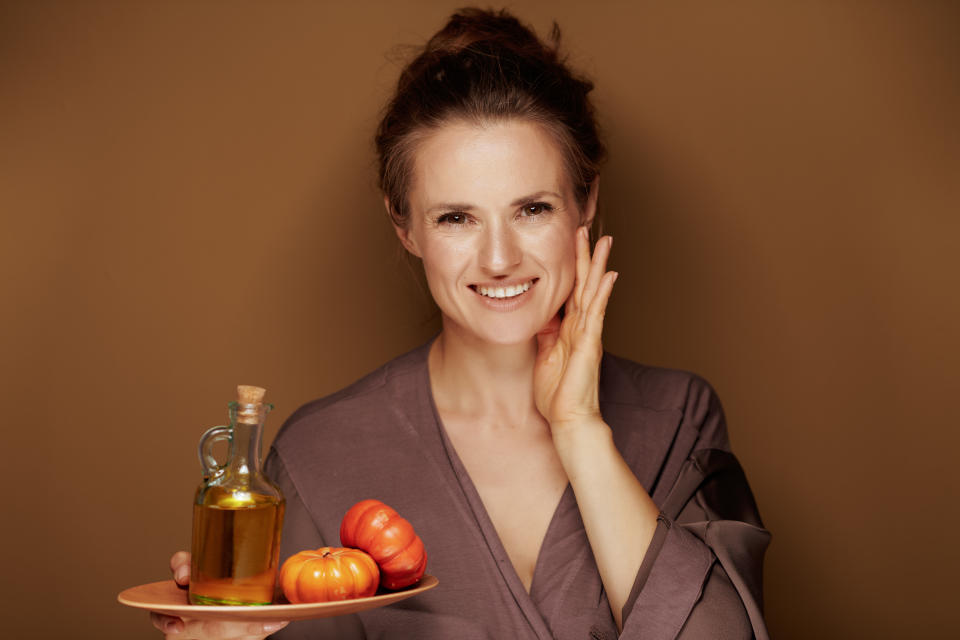 Hello autumn. Portrait of happy elegant middle age woman in a bathrobe showing pumpkin oil and touching face on bronze background.