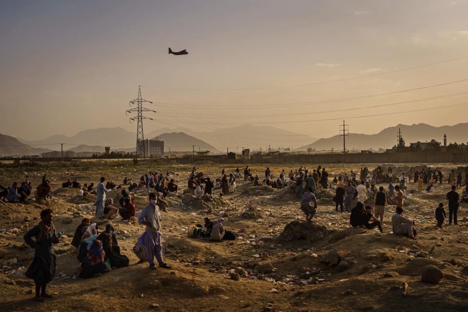 a helicopter flies over people standing in the hazy desert with mountains in the far distance