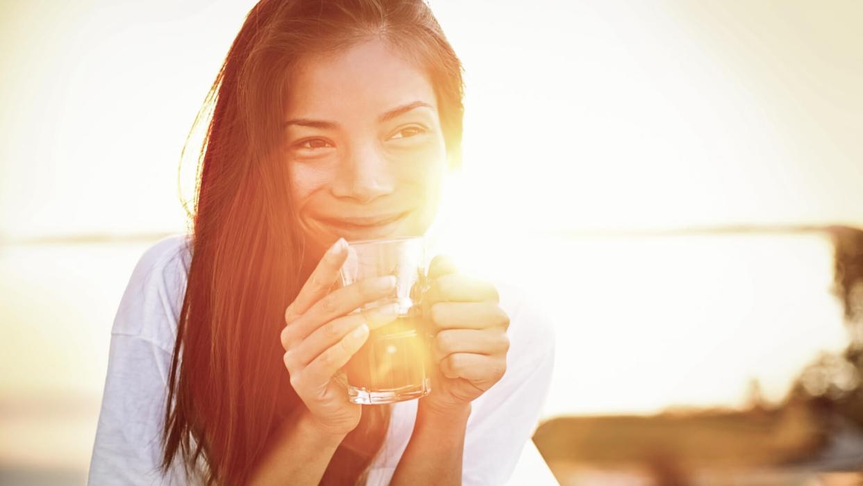 Asian woman drinking coffee in sun sitting outdoor in sunshine light enjoying her morning coffee.