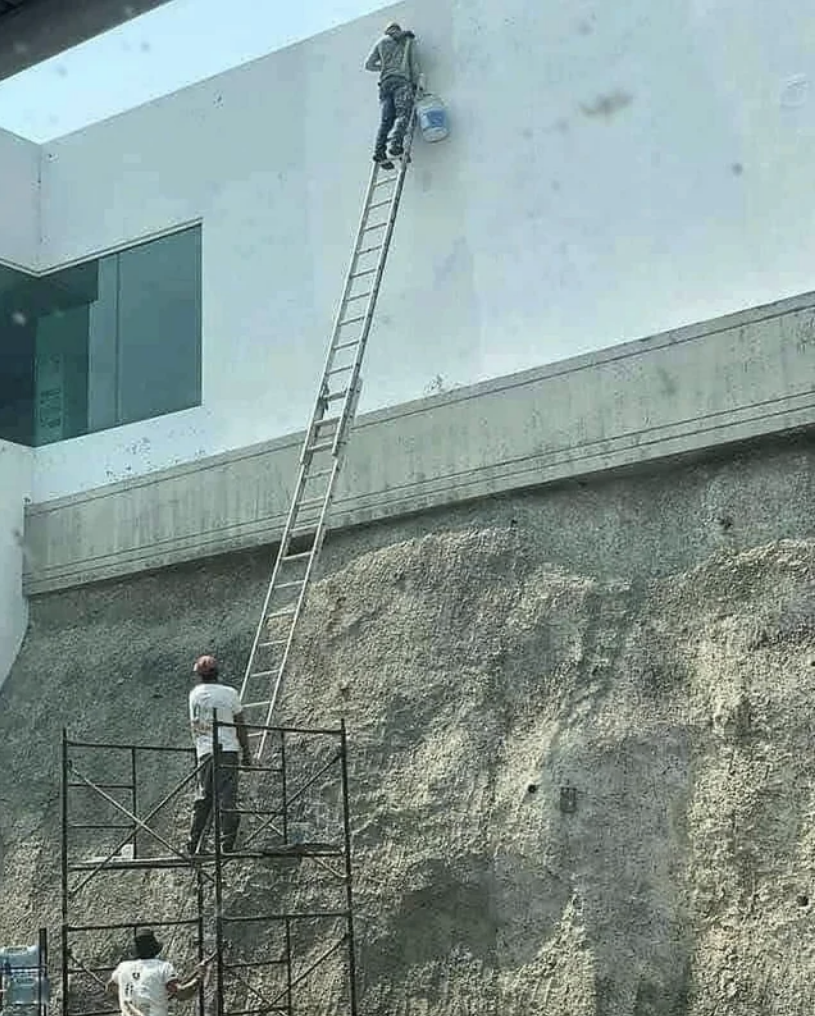 A ladder wasn't large enough to reach the top of a building, so one man is holding the bottom of the ladder off the ground while a second man climbs up it