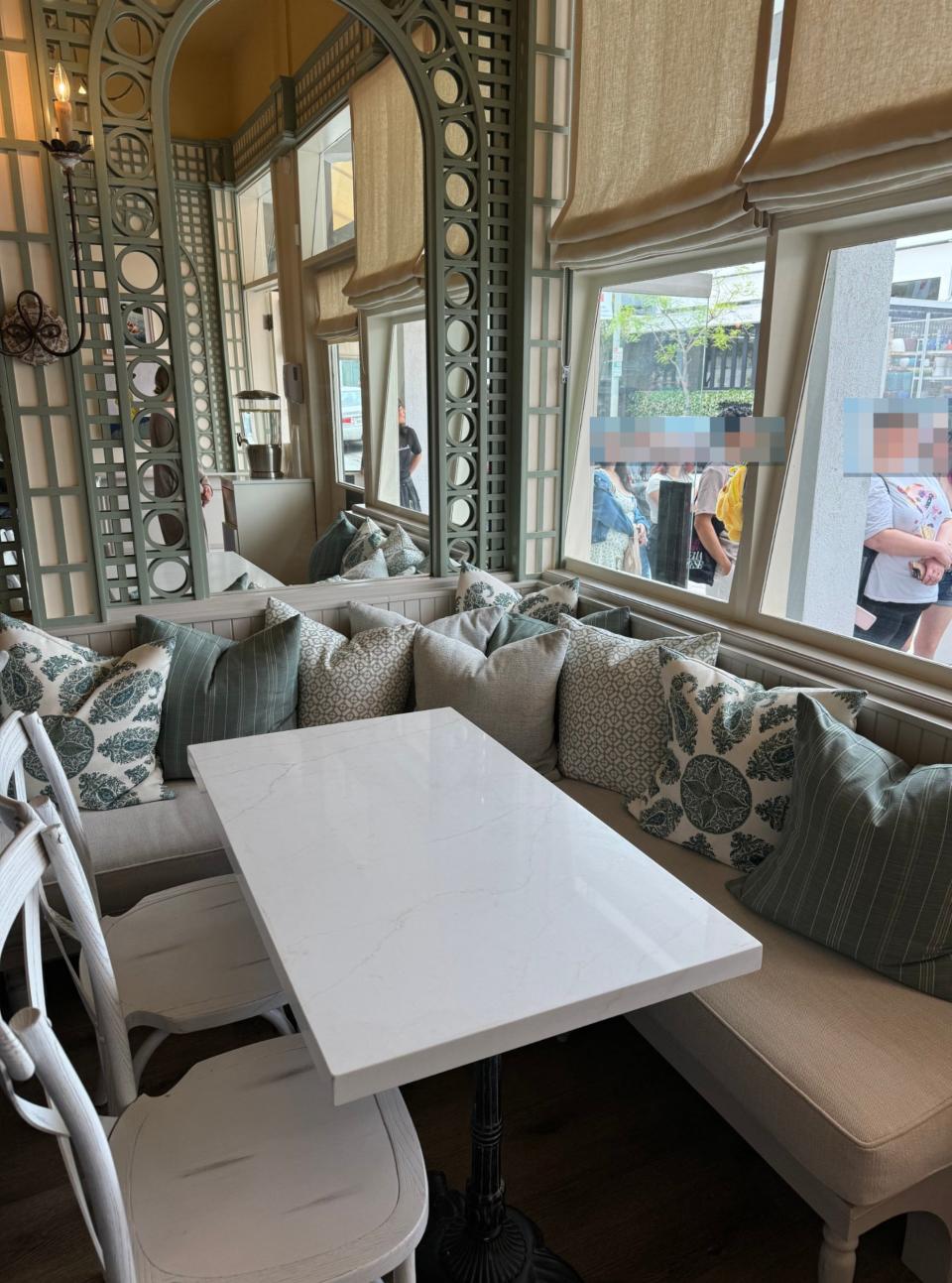 A cozy cafe corner with a white marble table, cushioned bench seating, decorative pillows, and large windows showing people standing outside