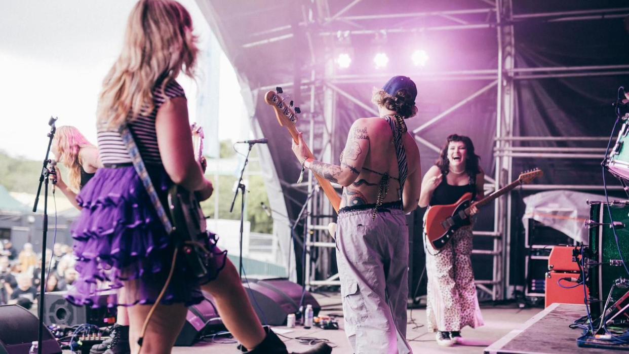 Panic Shack performing on stage at a festival. Megan, pictured on the right, smiles widely at her bandmates while holding her guitar. Bassist Emily Smith and guitarist Romi Lawrence are pictured with their backs to the camera while singer Sarah Harvey is at the top of the stage singing to the crowd. 