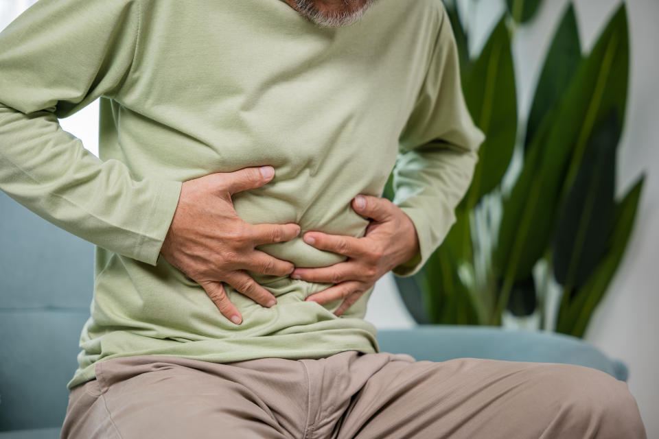 Elderly Asian old man sitting on sofa having stomachache, old age suffering from stomach ache holding his stomach in living room, people health problem, food poisoning