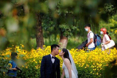 FILE - People wearing face masks pass by newlyweds kissing as they posing for wedding photos at the Olympic Forest Park in Beijing on July 2, 2020. Now that weddings have slowly cranked up under a patchwork of ever-shifting restrictions, horror stories from vendors are rolling in. Many are desperate to work after the coronavirus put an abrupt end to their incomes and feel compelled to put on their masks, grab their cameras and hope for the best. (AP Photo/Andy Wong)