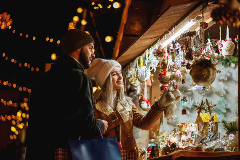 Happy smiling couple shopping at Christmas street market, choosing gifts. Winter holidays, vacation, travel, purchase conception. Outdoor night portrait.