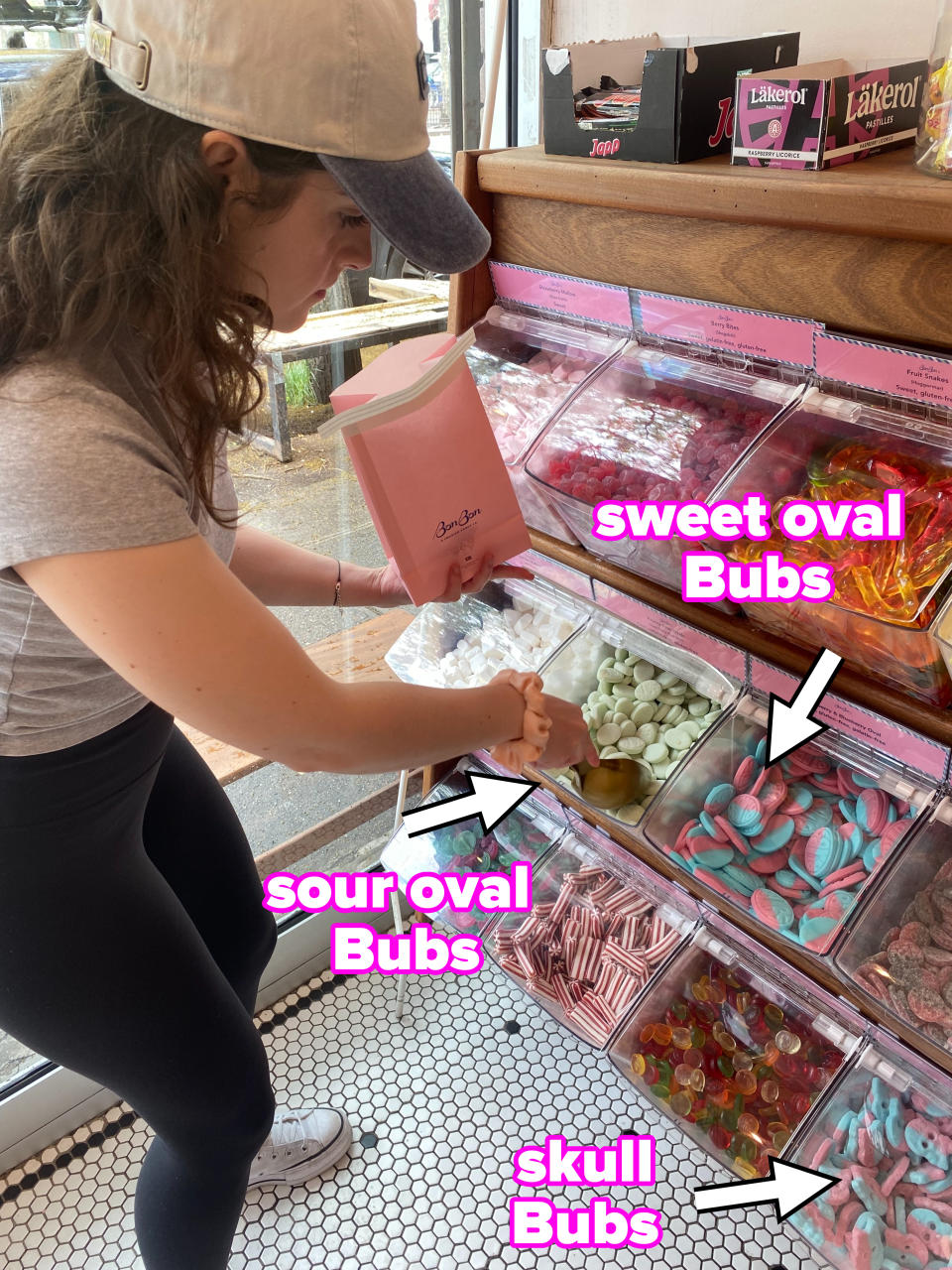 The author is scooping candies into a pink bag at a candy store. She is wearing a baseball cap, t-shirt, leggings, and sneakers