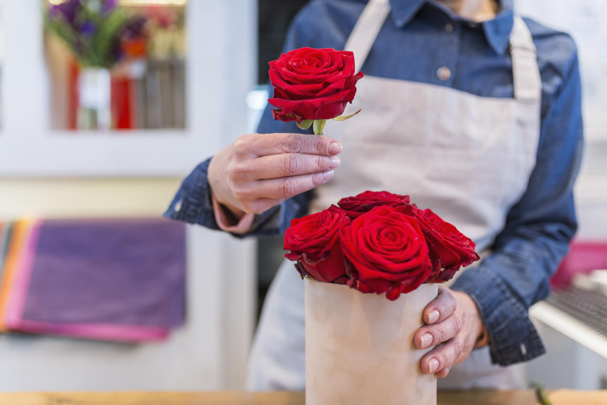 Photo of Female florist in flower shop or nursery presenting red roses, close-up. Female hands composing beautiful bouquet, close-up. Florist at work. Conceptual photo. Small business. Flowers delivery.  Florists creating order, making rose bouquet in flower shop wearing denim shirt and apron. 