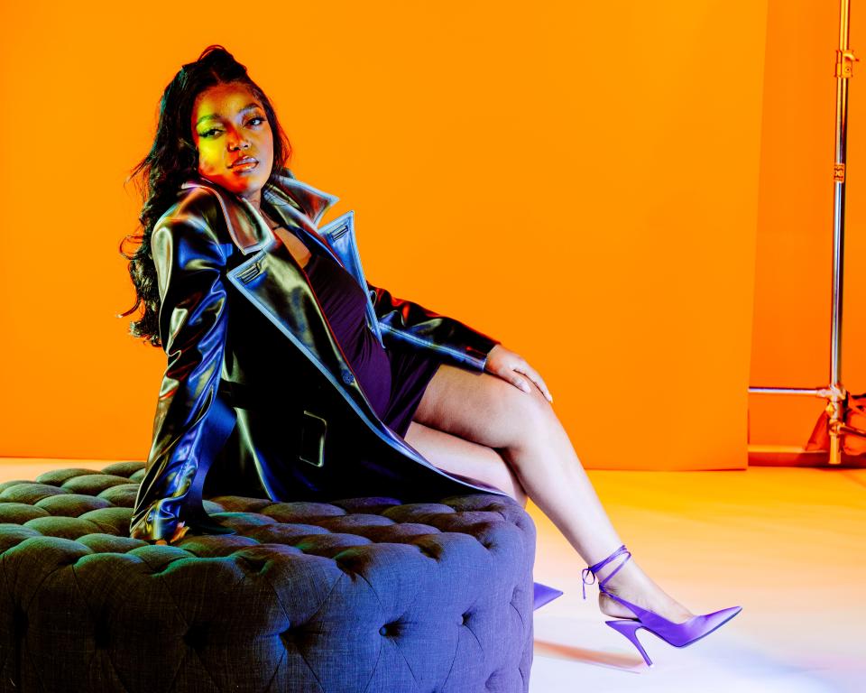 Anifa Mvuemba poses in a black leather coat and purple stiletto heels against a neon orange backdrop.