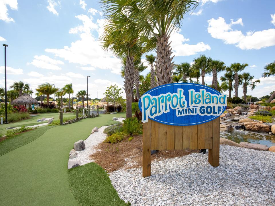 A sign that reads "parrot island mini golf" besides trees, turf.