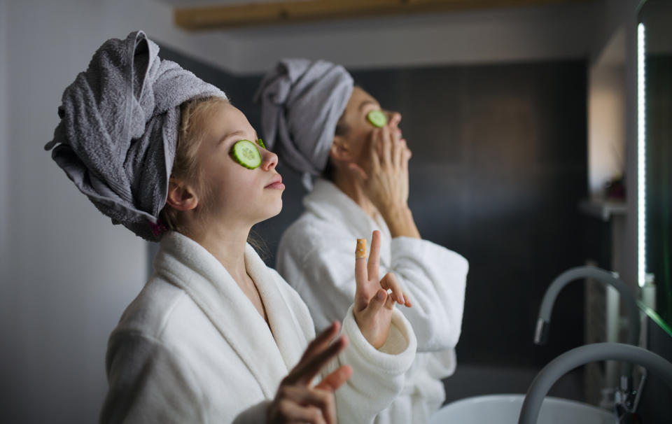 A parent and child enjoy a spa day at home, both wearing bathrobes and towels on their heads with cucumber slices on their eyes
