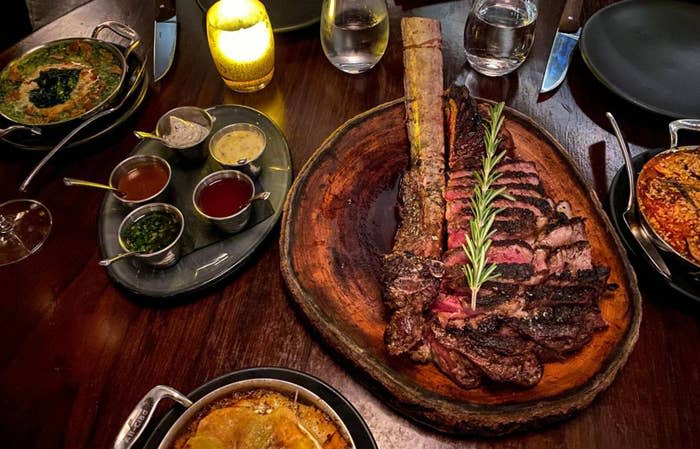 A large, bone-in steak garnished with rosemary on a wooden platter, surrounded by various sides and sauces, on a warmly lit dinner table.