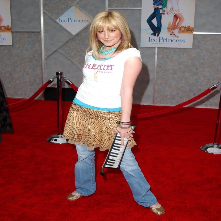 Ashley Tisdale wearing a graphic tee and skirt over jeans and a piano bag on a the red carpet for 