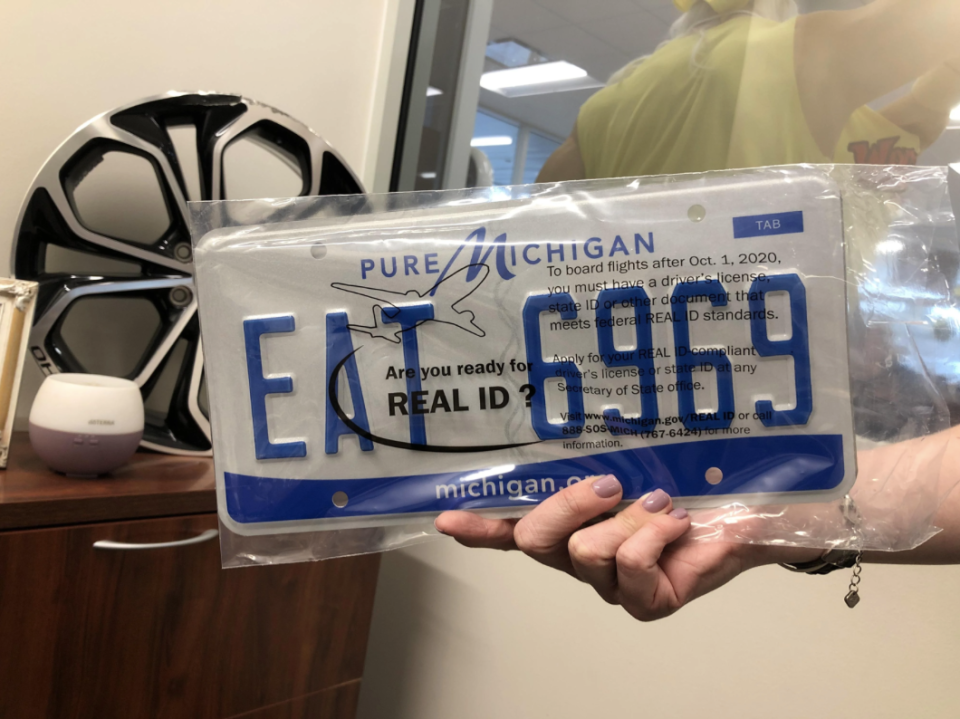 Person holding a Pure Michigan license plate that reads "EA7 6193," alongside a pamphlet. Items on the desk include a wheel rim and a small speaker