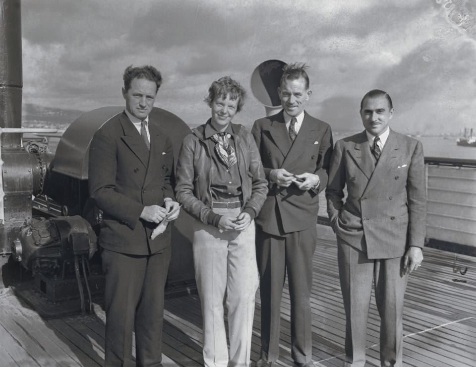 harry manning, amelia earhart, fred noonan, and paul mantz smile and stand on a ship deck, the men wear suits, earhart wears a leather jacket, collared shirt, scarf, and slacks