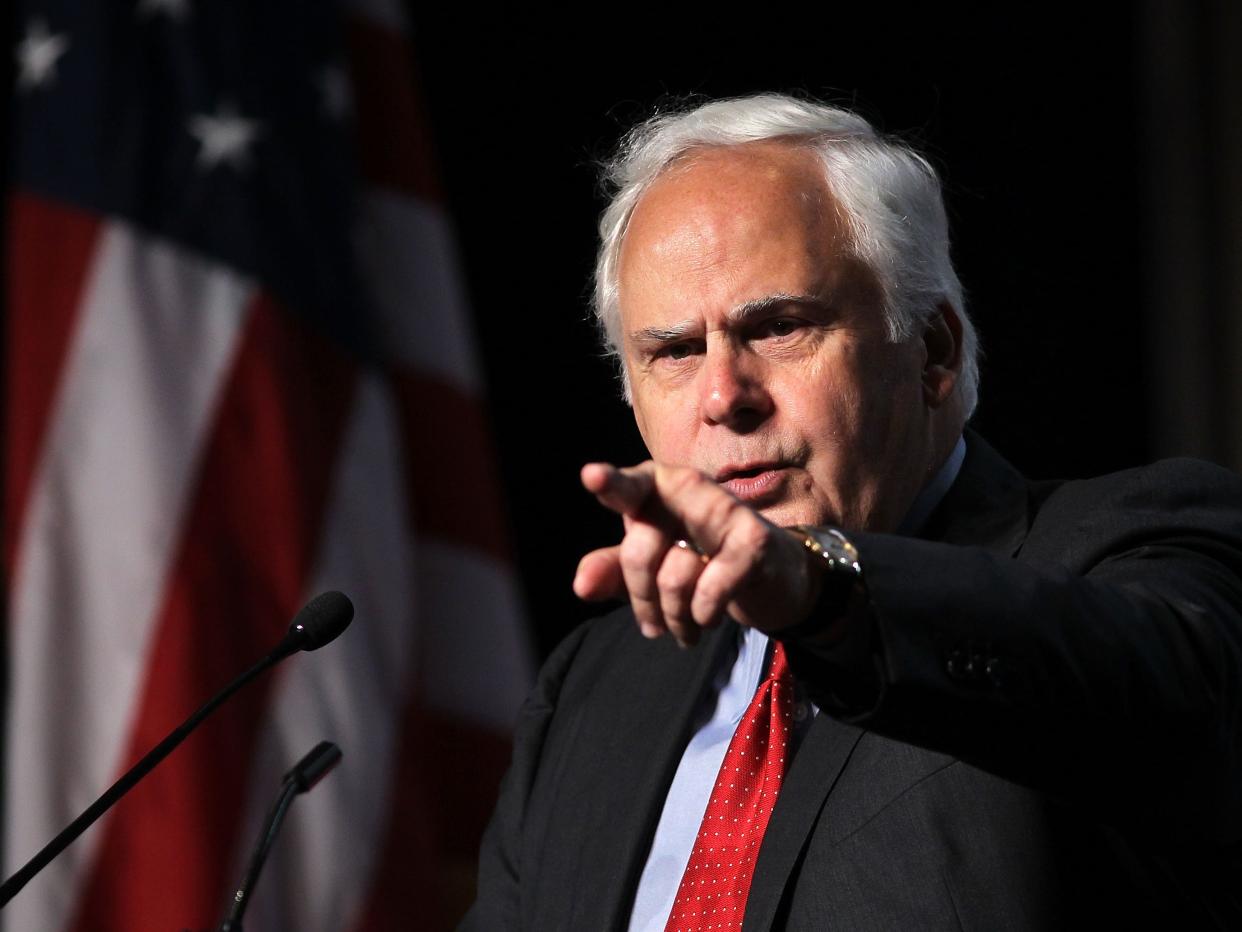 An older white man in a dark suit and red tie points at the camera in front of an American flag.