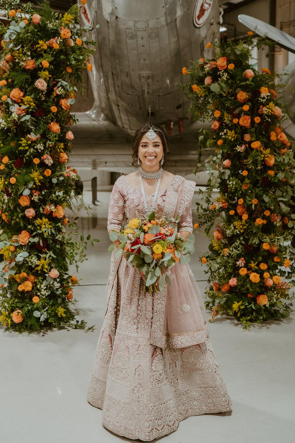 A bride stands in a purple lehenga in front of a floral archway and a plane.