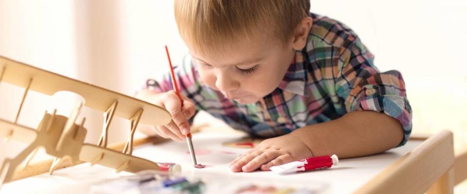 Adorable kid playing on his table with watercolors, painting a wooden parts of his airplane wooden toy. He is young designer with many ideas waiting to be found. Very shallow depth of field.