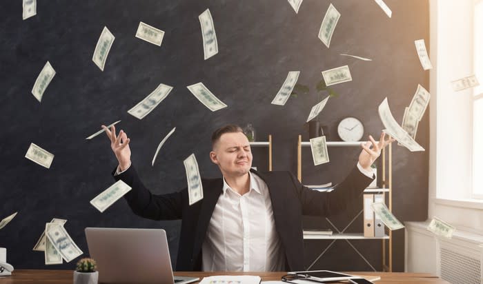 A man in a suit sits at a desk and poses with eyes closed and arms up as cash rains down on him.