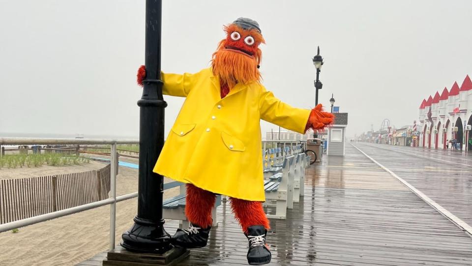 Gritty mascot wearing a yellow raincoat and hanging off of a black light pole on a beach boardwalk for his calendar
