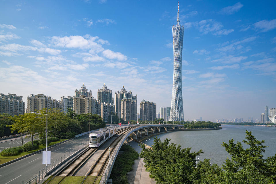A tram on tracks beside a river with the Canton Tower in the background and cityscape