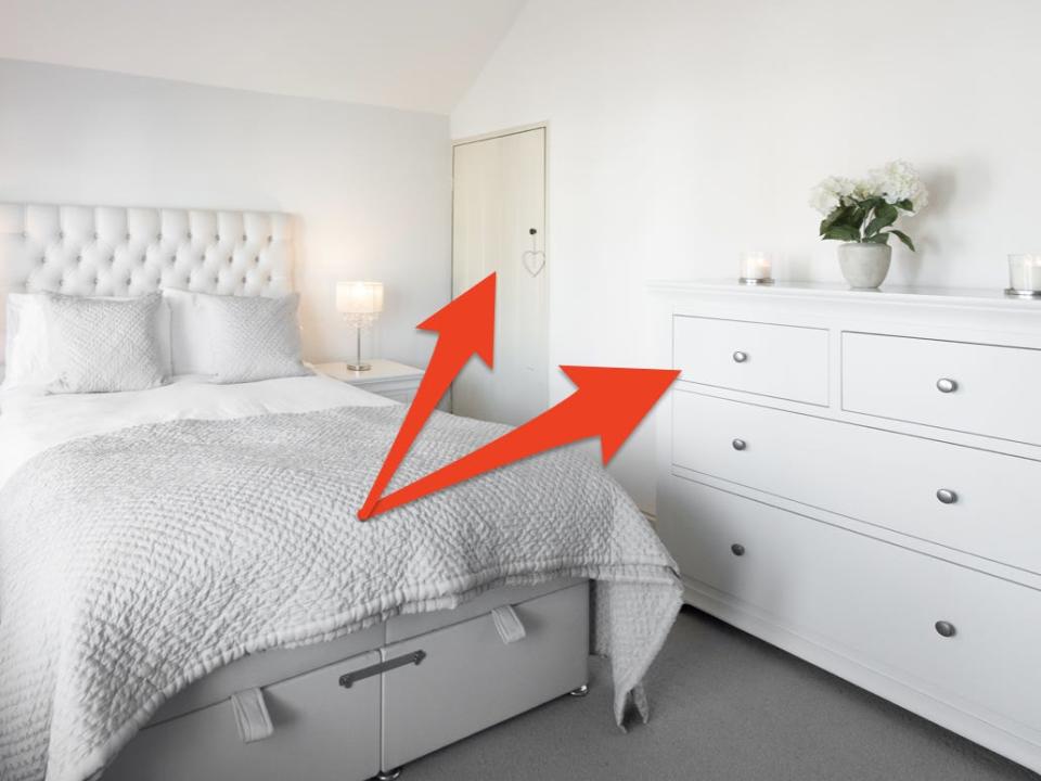 Bedroom with white sheets, white dresser, and white walls and door with red arrows pointing to dresser and door