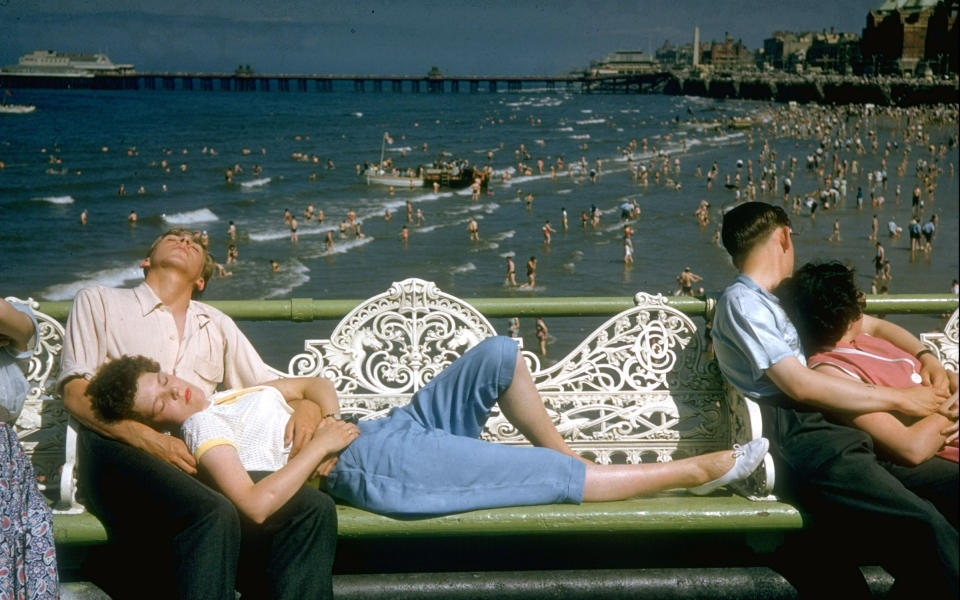 A couple dozes off on a wrought iron park bench in the United Kingdom in August 1955, while a busy beach scene continues in the backdrop.