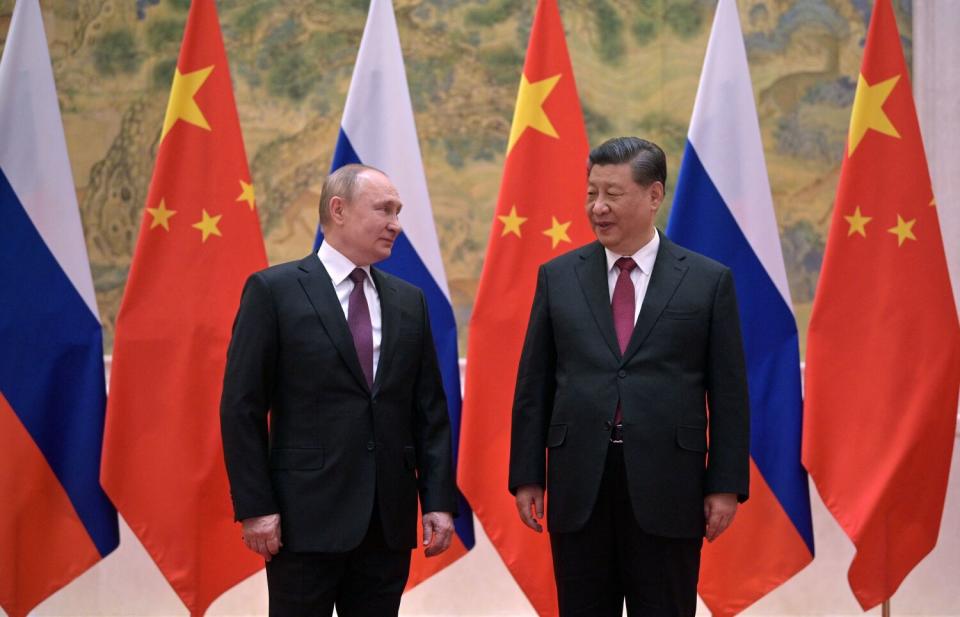 Two men in dark suits and red ties look at each other against a backdrop of red and tricolor flags