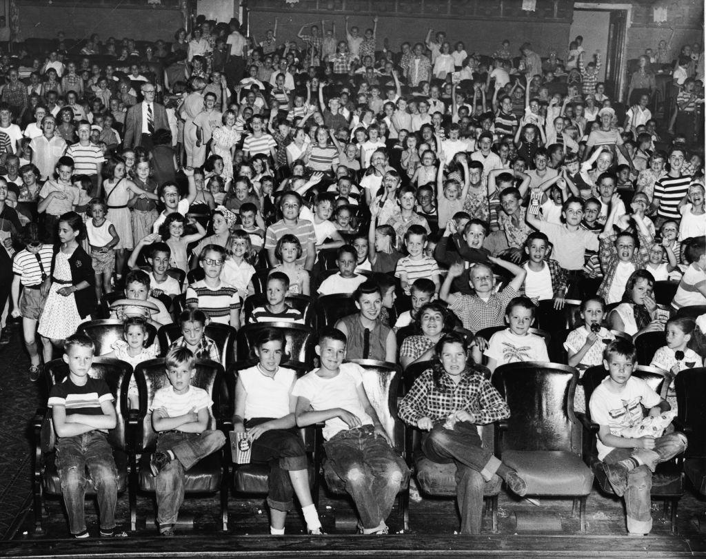 An audience of mostly children cheer and make faces in a movie theater, mid 1950s.