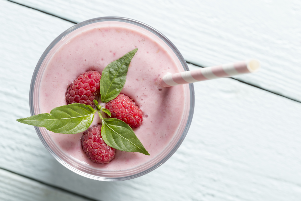 Top-view of a raspberry smoothie in a glass with a pink and white stripe straw with raspberries on top on a white wooden table